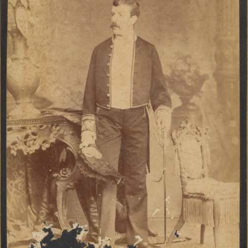 Undated photographic portrait of a man standing between a console table and a chair. He is holding a sword in his left hand a
