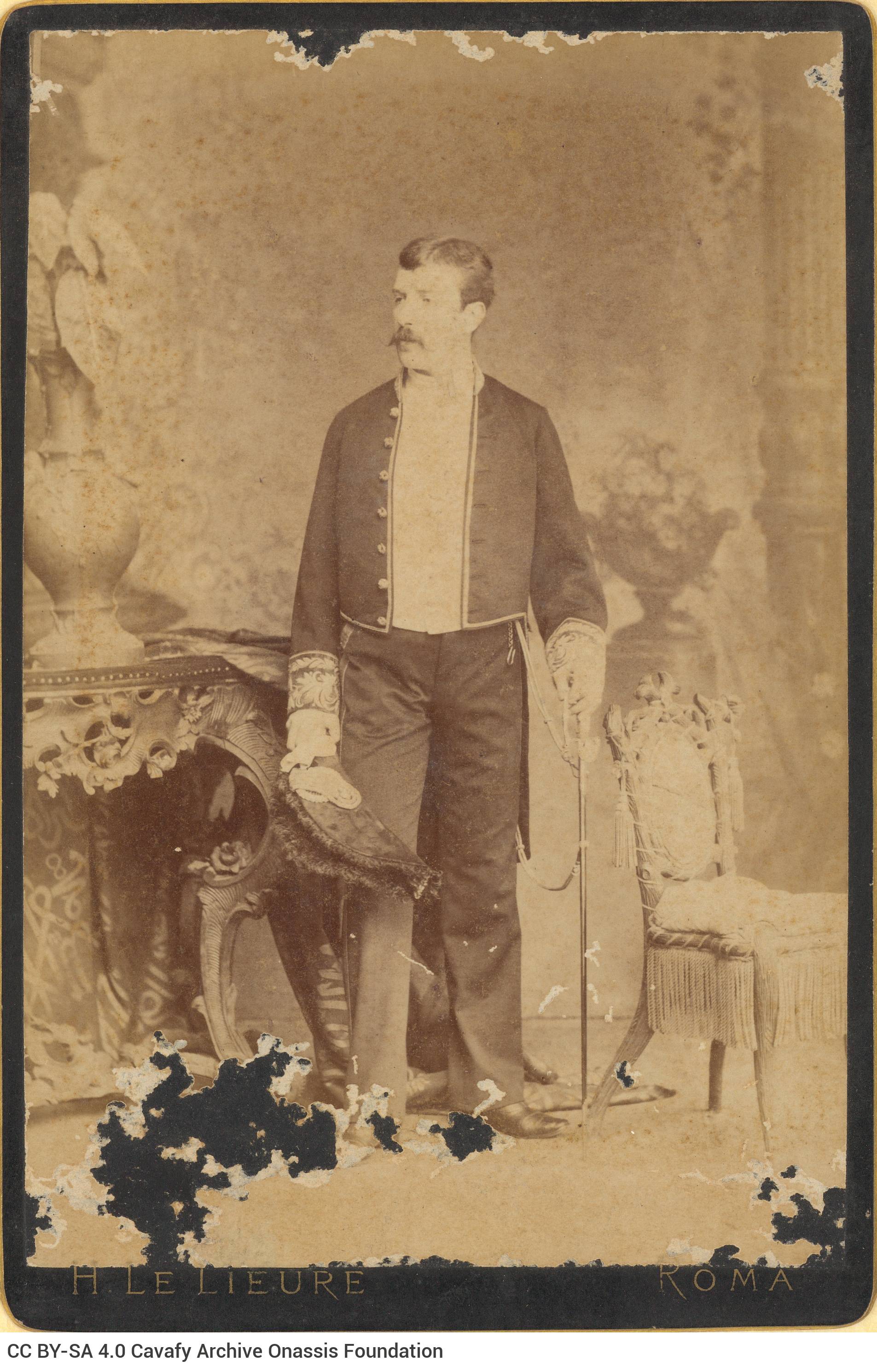Undated photographic portrait of a man standing between a console table and a chair. He is holding a sword in his left hand a