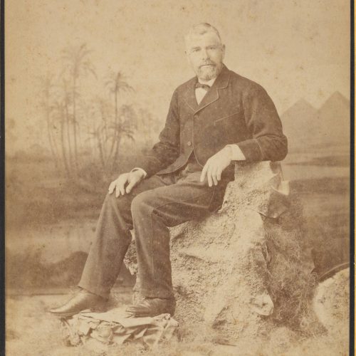 Photograph of a man in a suit and sitting on a rock, from the photo shop of N. Fettel & Cie of Alexandria. The photo shop's n