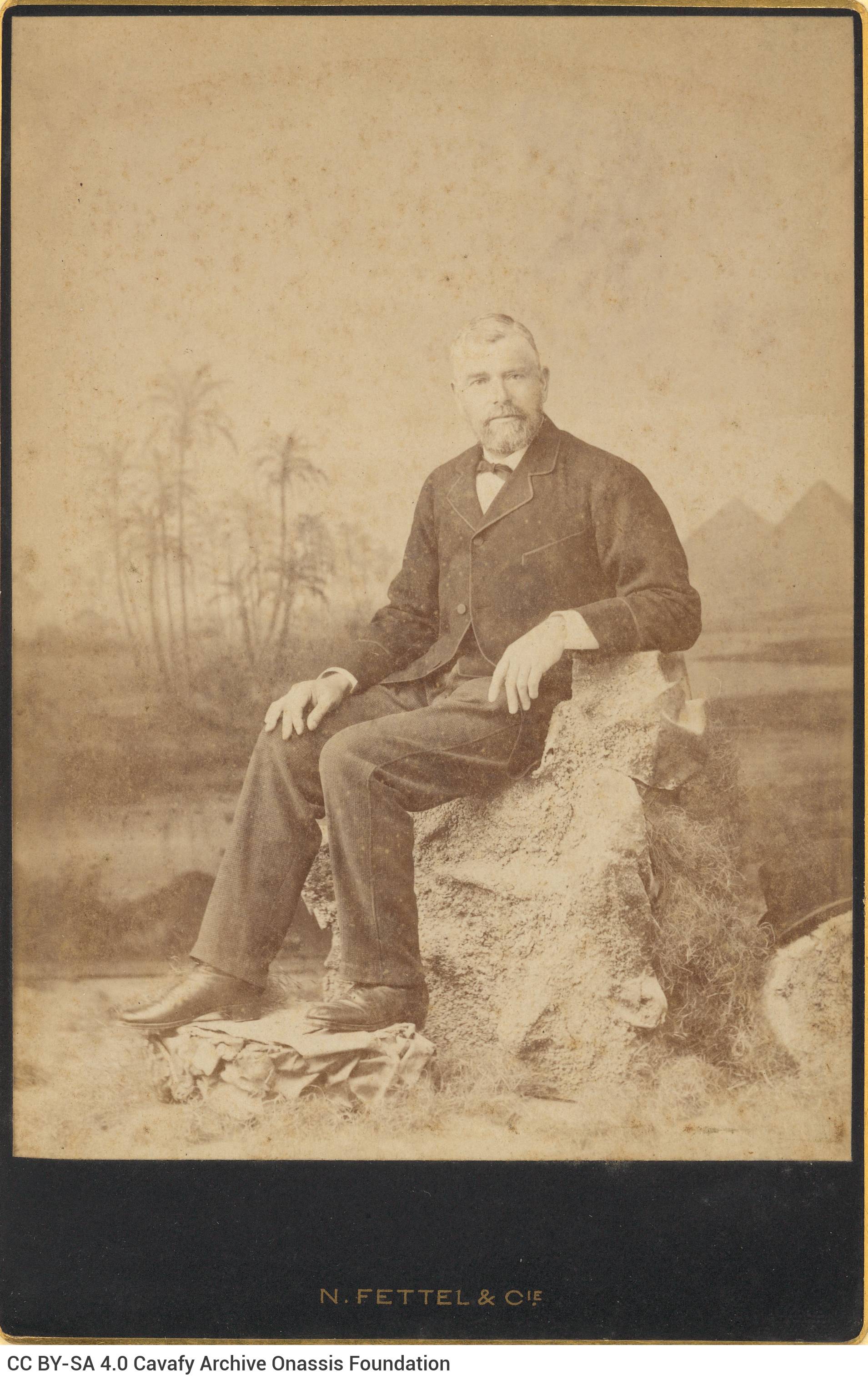 Photograph of a man in a suit and sitting on a rock, from the photo shop of N. Fettel & Cie of Alexandria. The photo shop's n