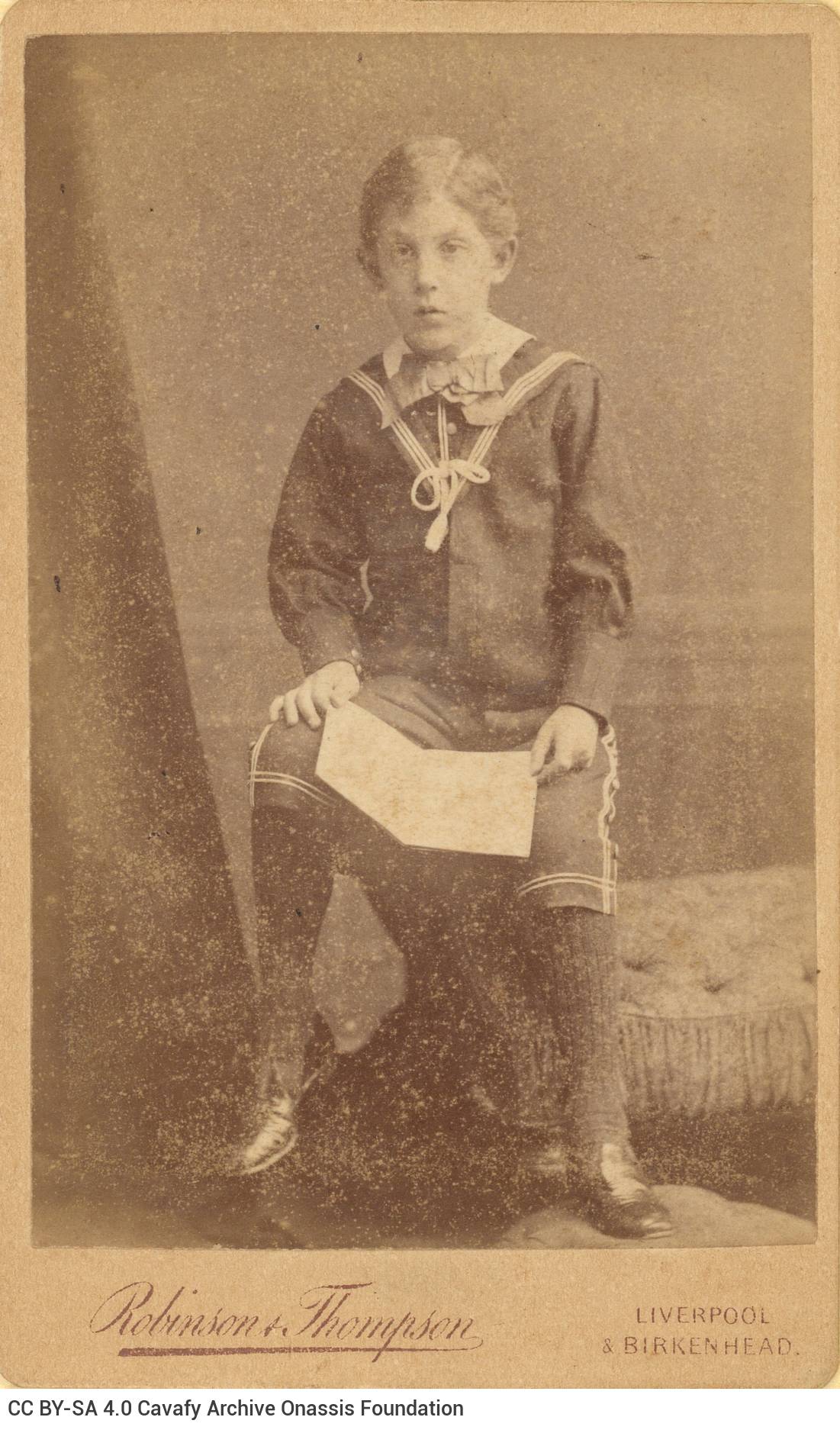 Photograph of a boy in sailor's clothes, sitting on the arm of a seat and holding a book in his hands. The logo of the photo 