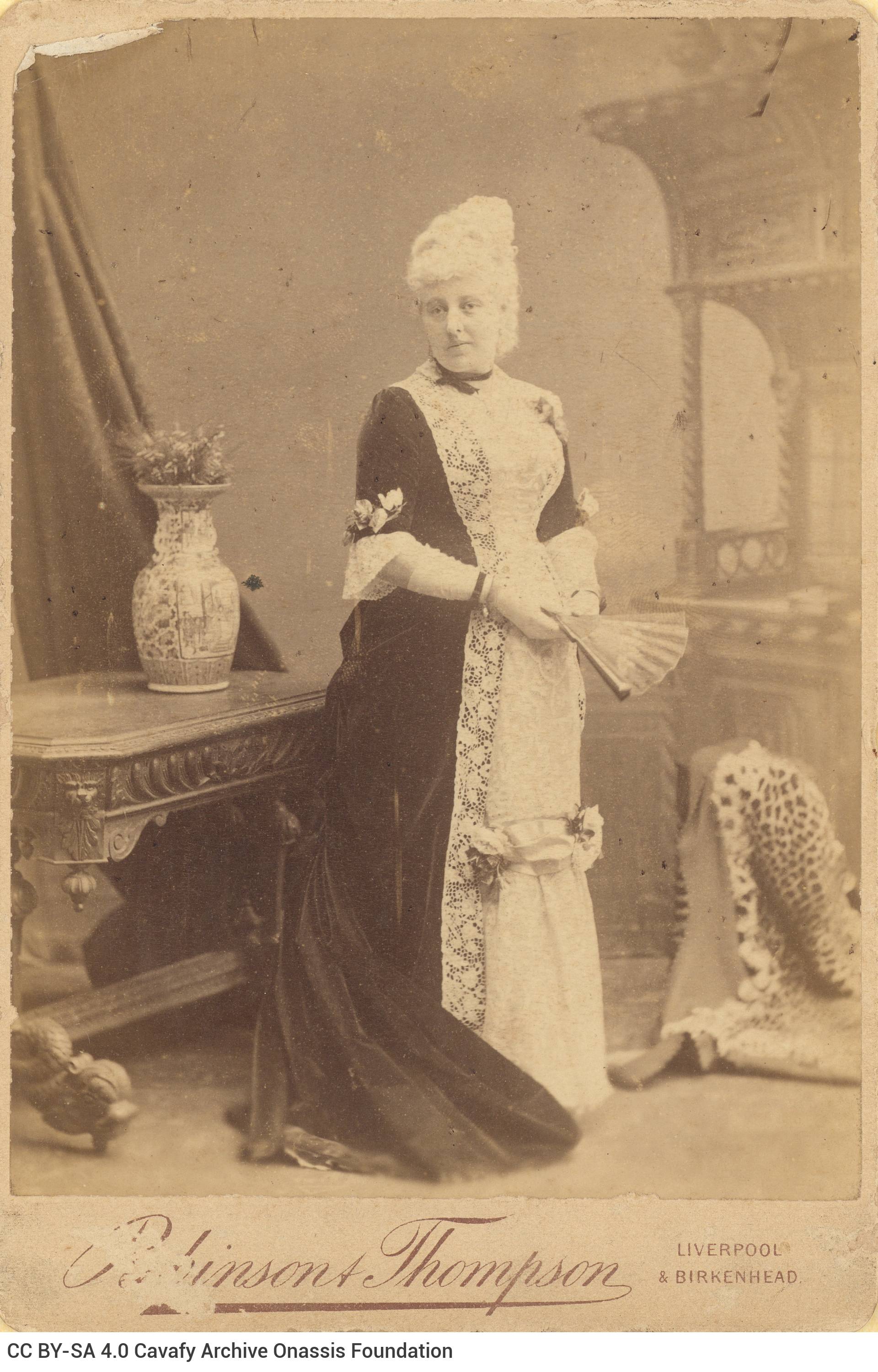 Photograph of a woman standing next to a table. She is wearing a long dress with a tail and is holding a fan. Handwritten dat