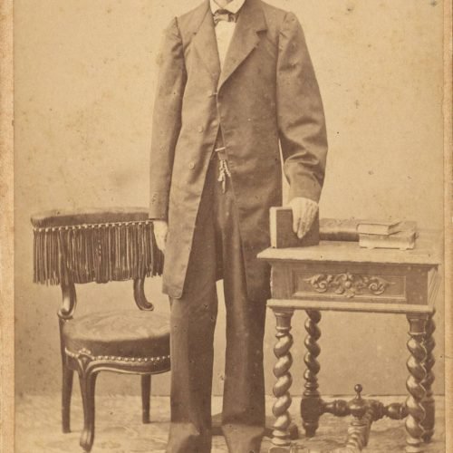 Photograph of the poet's father, Peter John Cavafy. He is depicted standing between a seat and a table, with a book in his le
