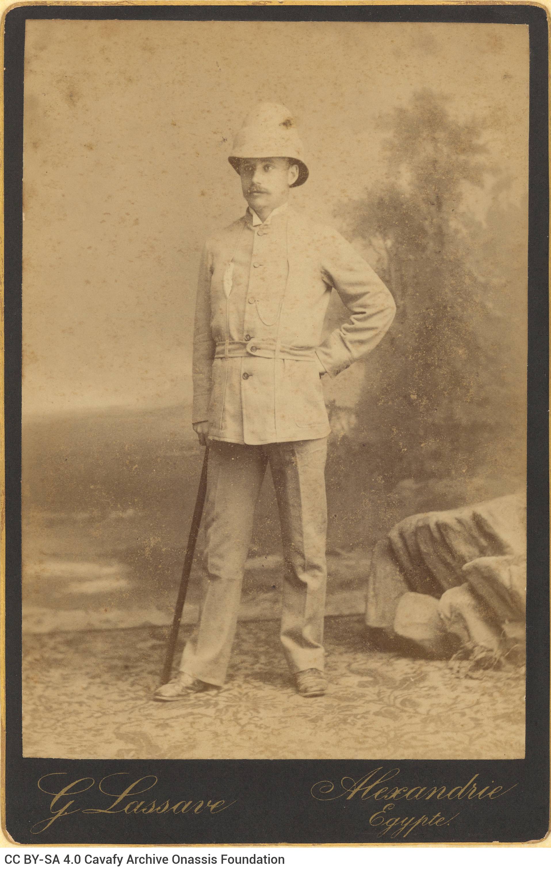 Photograph of a standing man in colonial apparel and a colonial hat. He is holding a cane in his right hand. The logo of the 