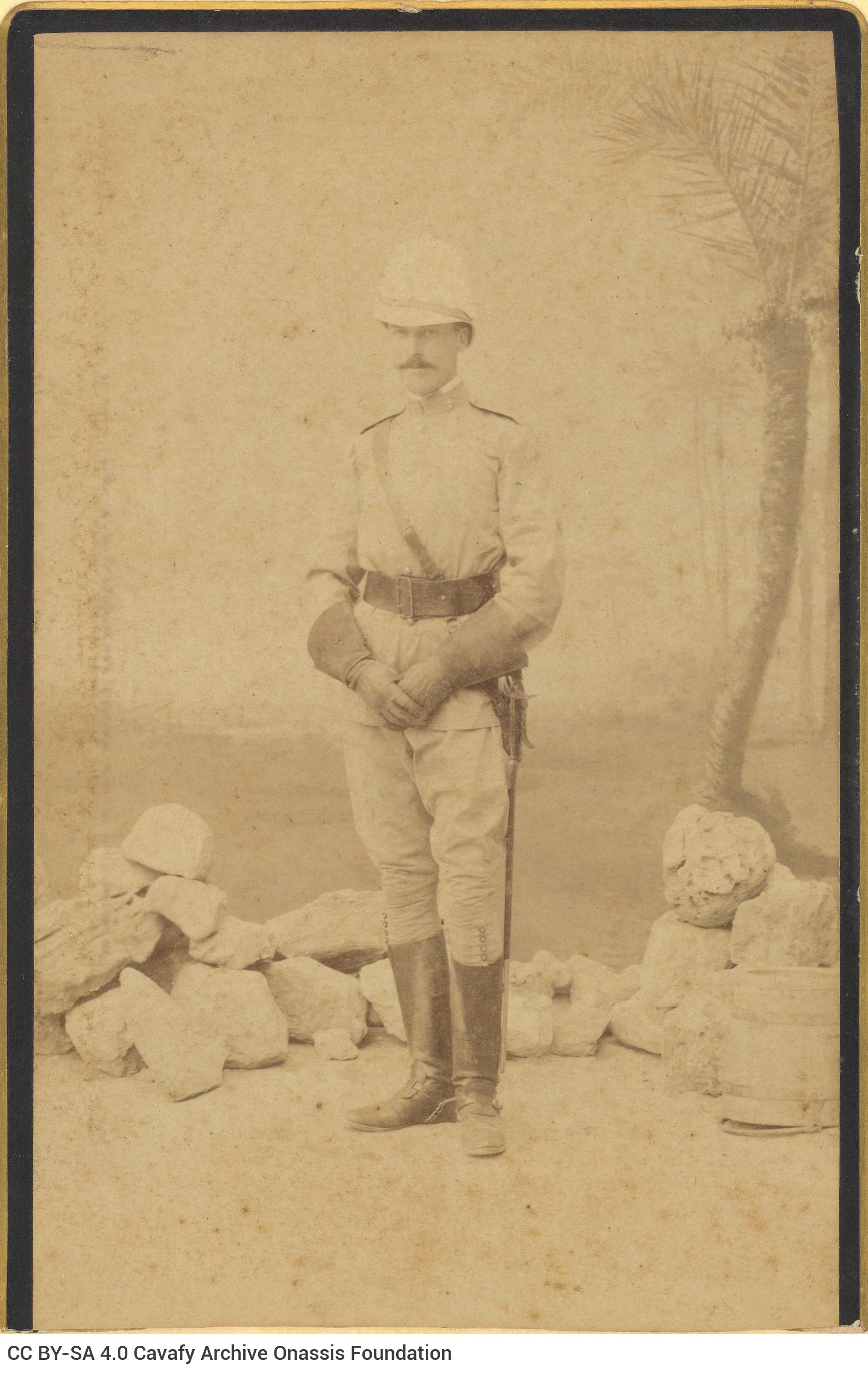 Photograph portrait of a standing man in a military uniform and colonial hat. He is wearing boots and gloves. The lower part 