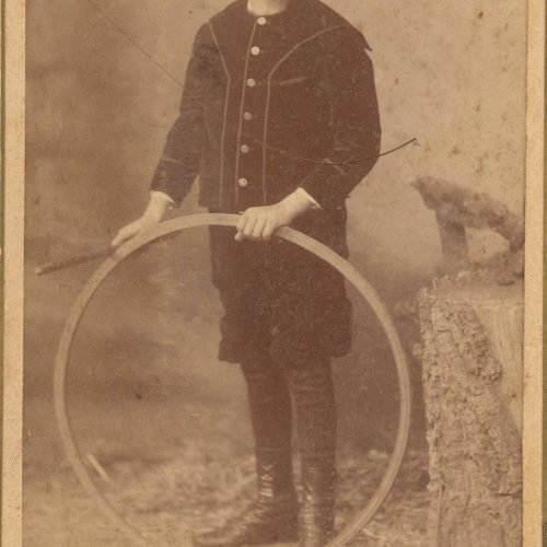 Φωτογραφία αγοριού με σκουρόχρωμα ρούχα. Κρατάει στεφάνι και ραβδί. 