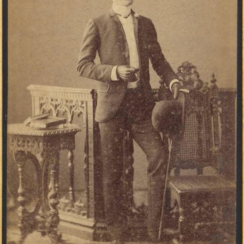 Photographic portrait of a young man with a moustache and leather gloves, standing between a table and a chair. He is holding